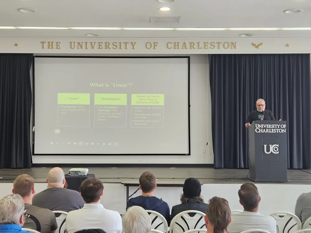 A speaker at the University of Charleston podium in the GSU Ballroom, presenting a slide titled What is Linux to a seated audience